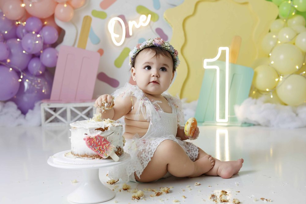 Baby holding a donut and a handful of cake beside a frosted smash cake on a white stand