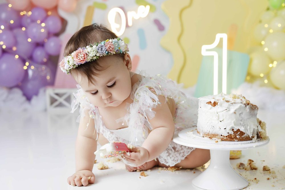 Baby in a flower crown leaning forward with a cookie popsicle next to a half-eaten smash cake