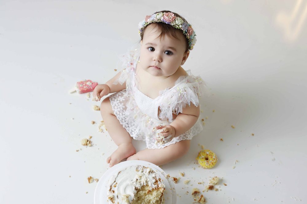 Overhead view of a baby in a lace romper sitting among cake crumbs and a donut, looking up at the camera