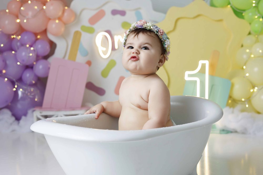 Baby pursing her lips in a white clawfoot tub after her cake smash, neon '1' and pastel balloons behind