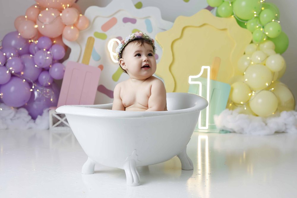 Baby smiling upward in a small white clawfoot tub between purple, peach and green balloon garlands