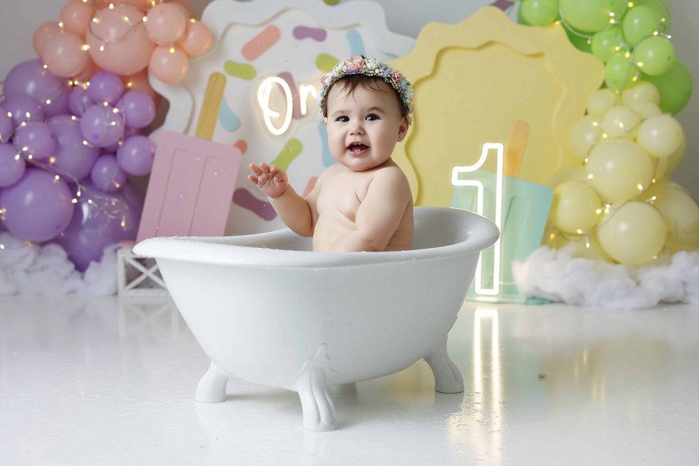 Baby waving from a white clawfoot tub on a pastel popsicle set with a glowing neon 'one' sign