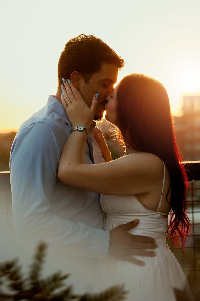 Couple kissing in golden backlight, her hands cupping his face, city rooftop softly blurred behind them