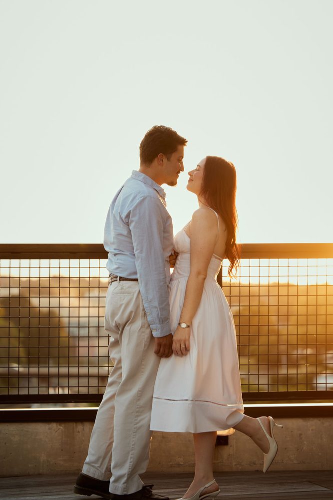 Couple in a light blue shirt and white dress smiling nose to nose against a rooftop railing at golden hour