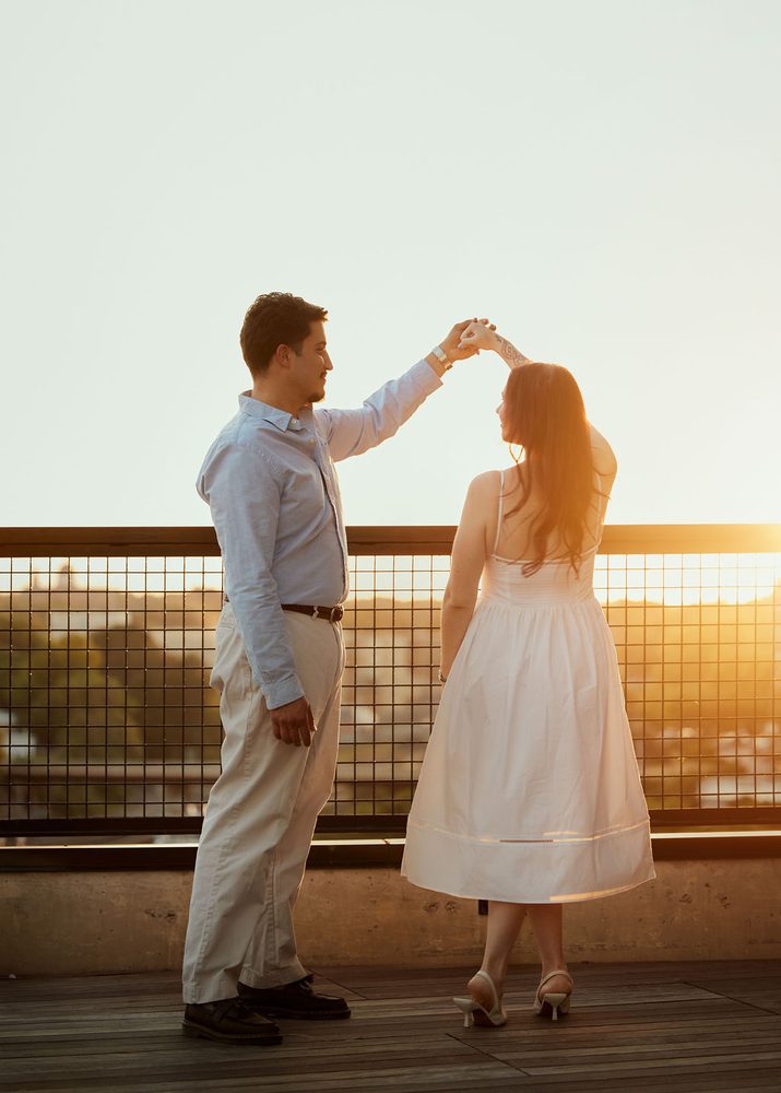 Man twirling his partner in a white sundress on a rooftop deck overlooking trees at sunset