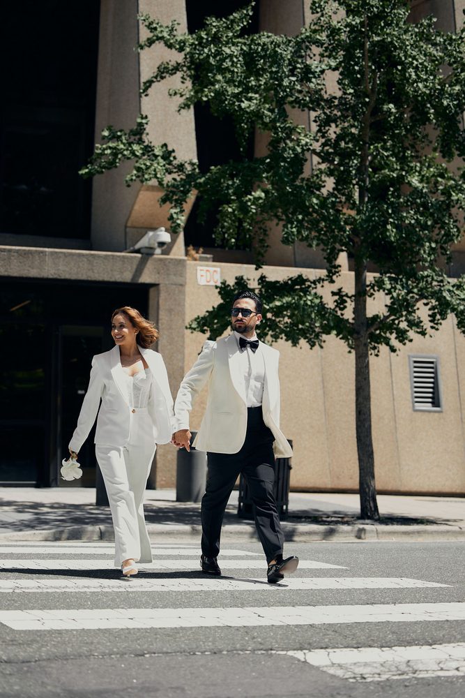 Newlyweds in a white pantsuit and ivory tuxedo jacket walking hand in hand across a sunny city crosswalk
