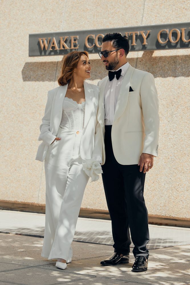 Bride and groom smiling at each other beneath the Wake County courthouse sign on a sunlit sidewalk