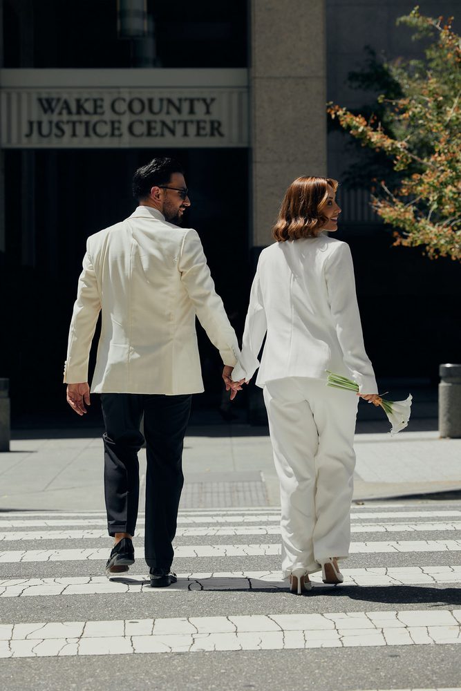 Newlyweds holding hands on the crosswalk, the bride smiling back at her groom under the justice center sign