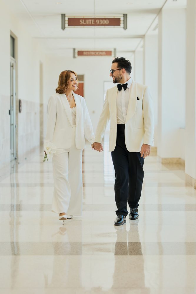 Wide shot of the bride and groom strolling a long marble hallway beneath a Suite 9300 sign