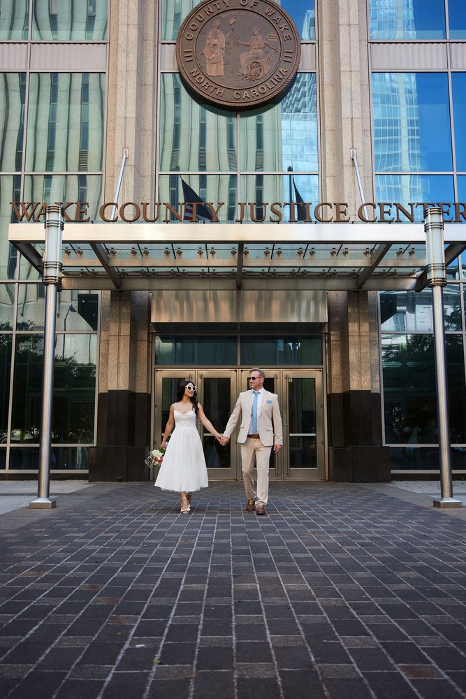 Bride in a tea-length white dress and groom in a tan suit walking out of the Wake County Justice Center