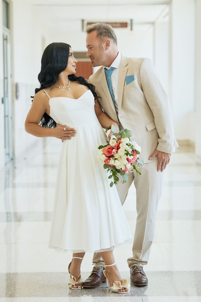 Bride with a pink and white rose bouquet gazing at her groom in a tan suit and blue tie in a courthouse hallway