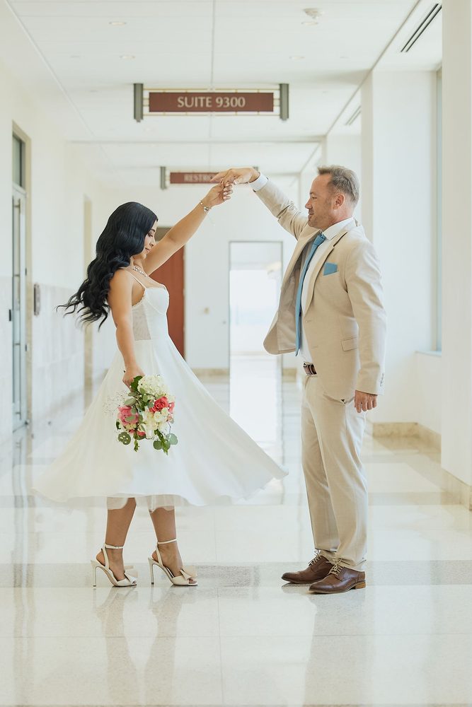 Groom twirls the bride in her flowing white midi dress in a gleaming marble courthouse corridor