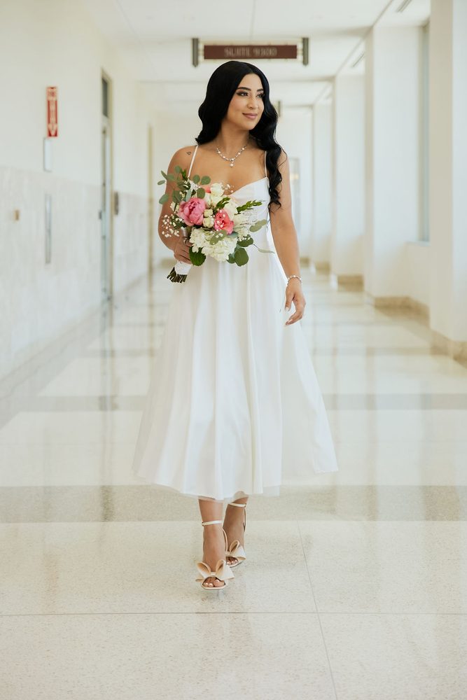 Bride walking alone down a courthouse hallway with a peony and rose bouquet and bow-detail heels