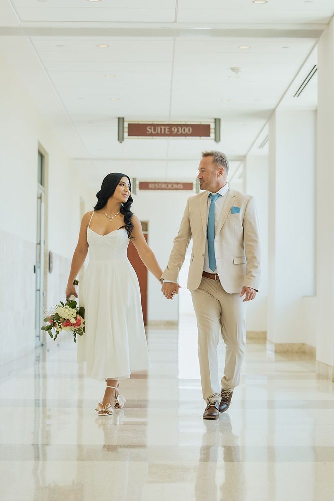 Couple holding hands down a bright courthouse hallway, the bride glancing up at her groom in a tan suit