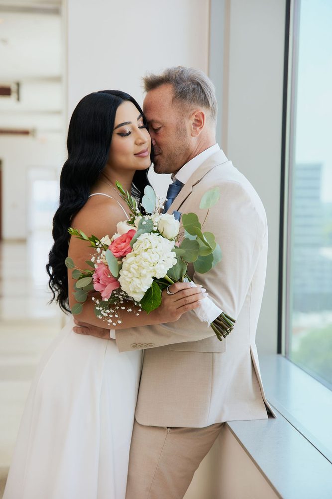 Groom nuzzles the bride's temple by a tall window as she holds her bouquet of roses and hydrangeas