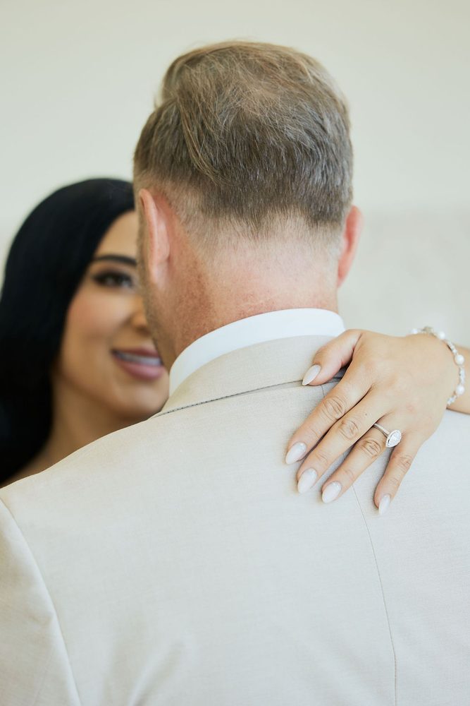Bride's hand with her engagement ring resting on the groom's shoulder as she smiles during an embrace