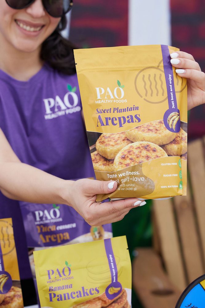 Smiling vendor in a purple shirt holding up a bag of sweet plantain arepas at a food brand booth