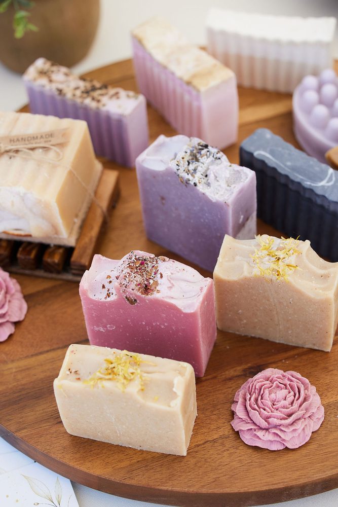 Handmade soap bars in lavender, pink and cream topped with dried petals on a round wooden tray