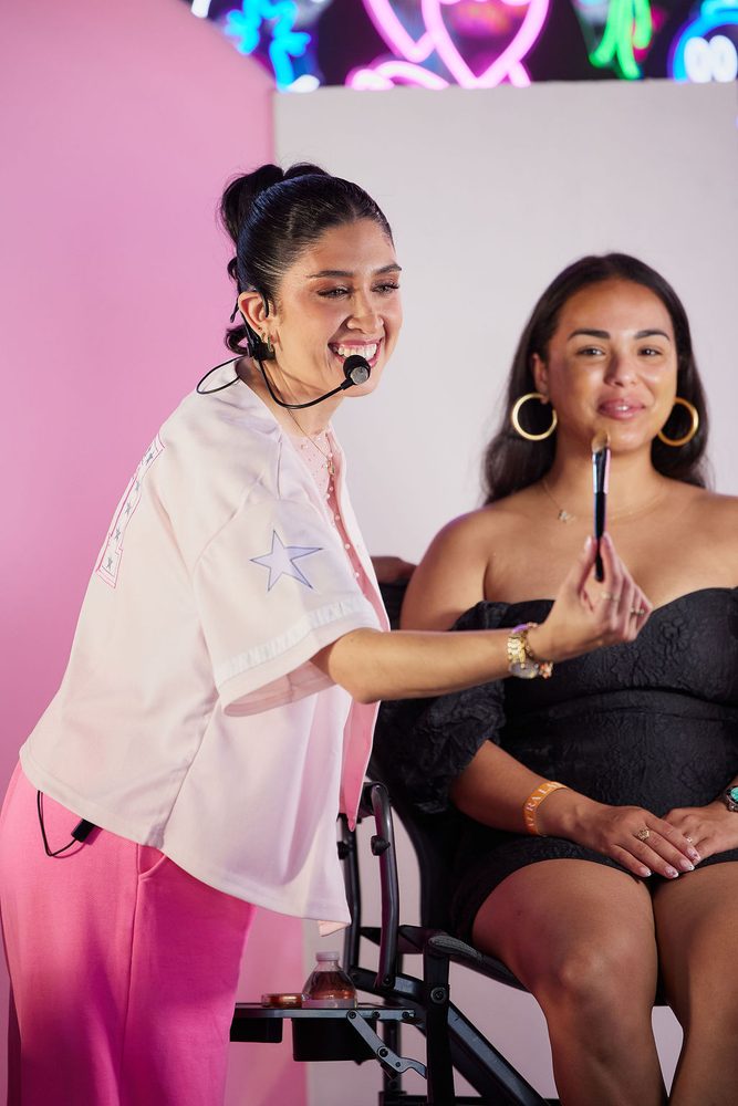 Smiling makeup artist with a headset mic holding a brush up beside a seated model in a black dress