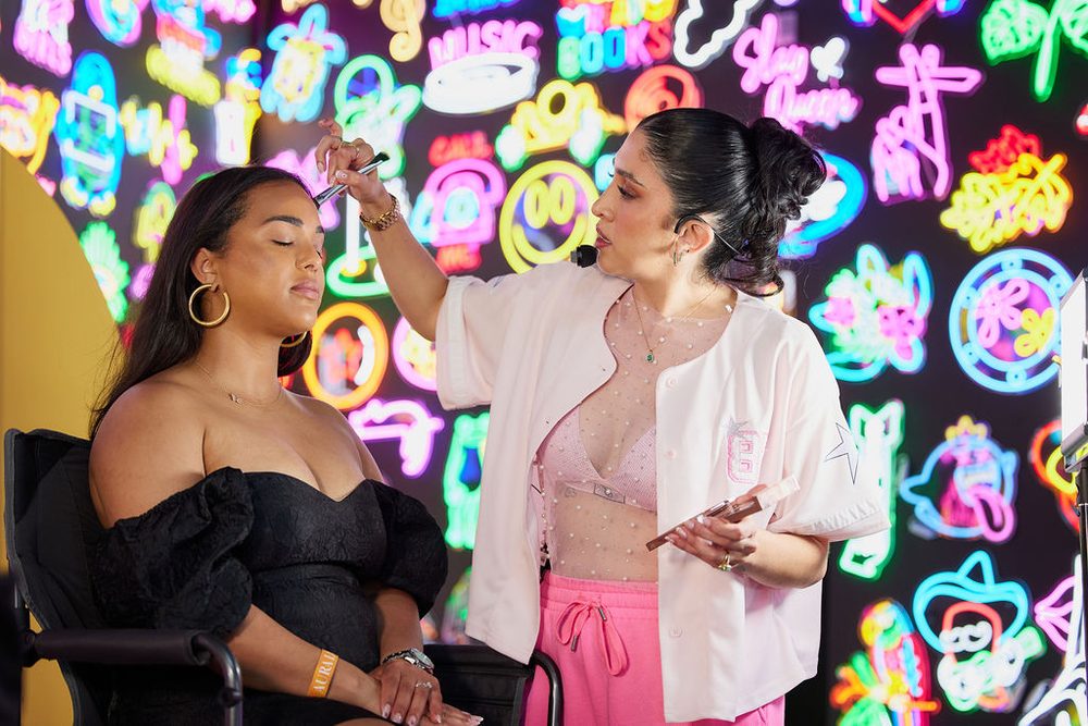 Makeup artist sweeping a brush across a seated model's face against a bright wall of neon signs