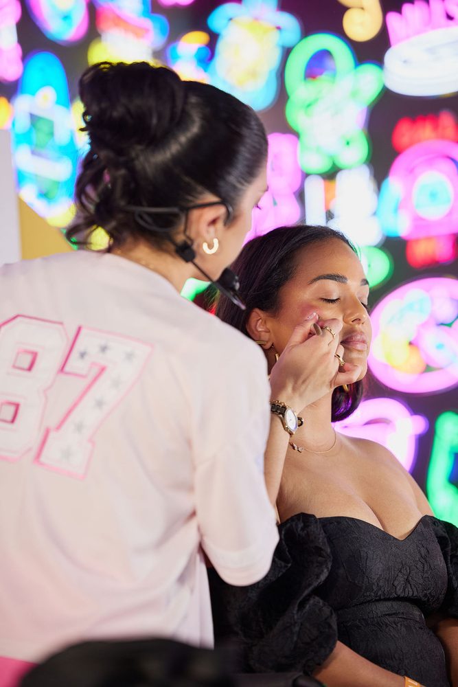 Over-the-shoulder view of a makeup artist blending blush on a model's cheek under colorful neon lights