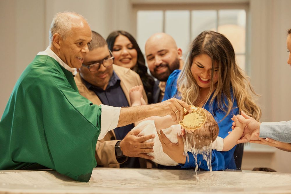 Officiant in green vestments pouring water over a baby's head at a baptismal font as smiling family looks on