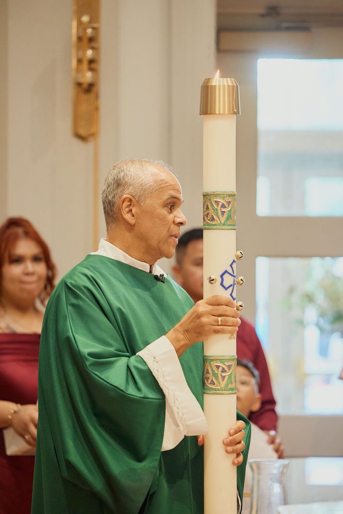 Officiant in green vestments holding a tall decorated candle during a baptism ceremony in a bright chapel