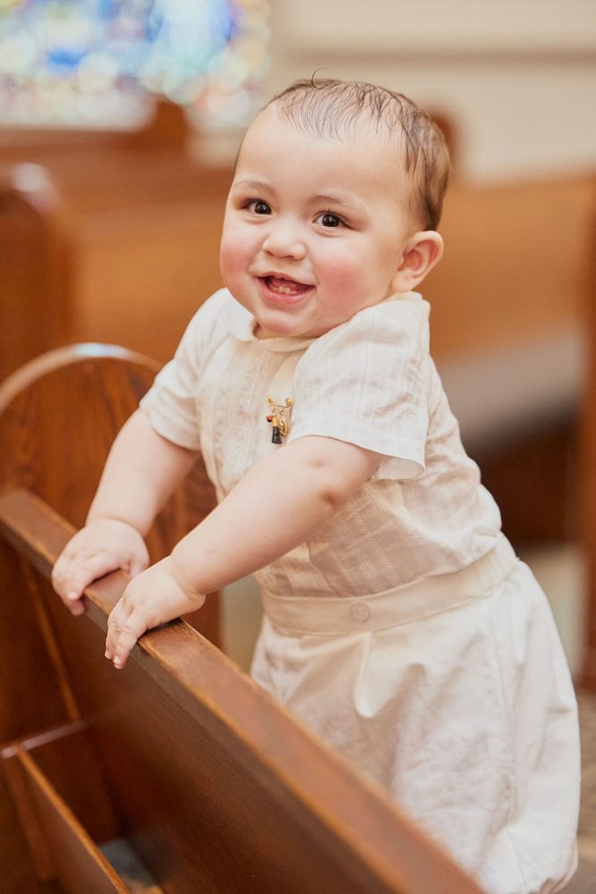 Smiling baby in a cream christening outfit standing and holding onto a wooden church pew