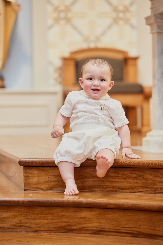 Barefoot baby in a white christening romper sitting on polished wooden steps and smiling at the camera