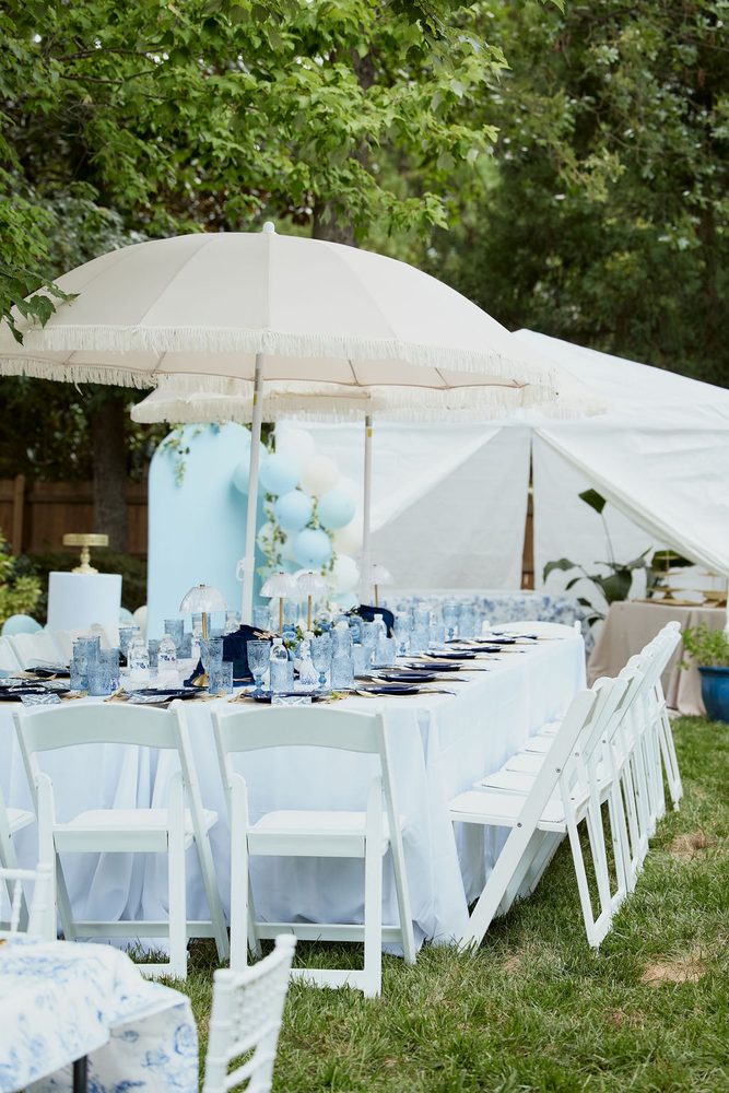 Long reception table with blue glassware under fringed white umbrellas, balloons and a tent in a backyard