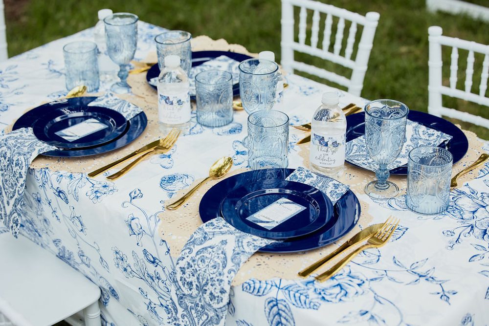 Place settings with navy plates, gold flatware and blue glassware on a blue-and-white floral tablecloth