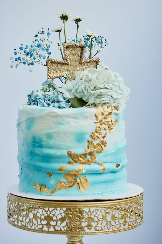 Blue watercolor cake with gold leaf, a gold cross topper and fresh flowers on an ornate gold stand