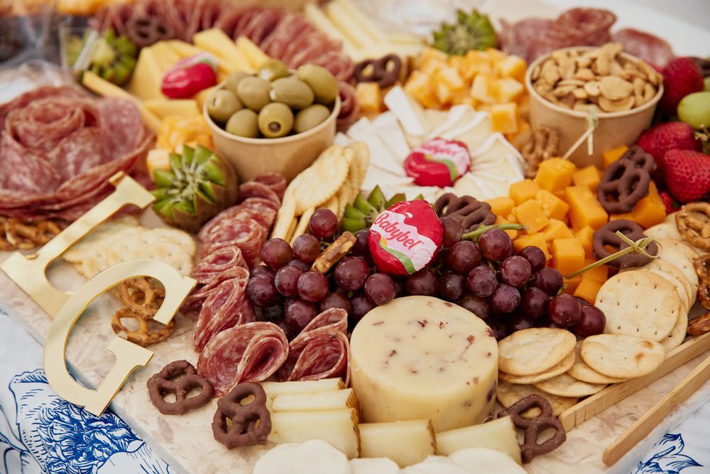 Charcuterie board of salami roses, cheeses, grapes, olives, crackers and chocolate pretzels with a gold letter