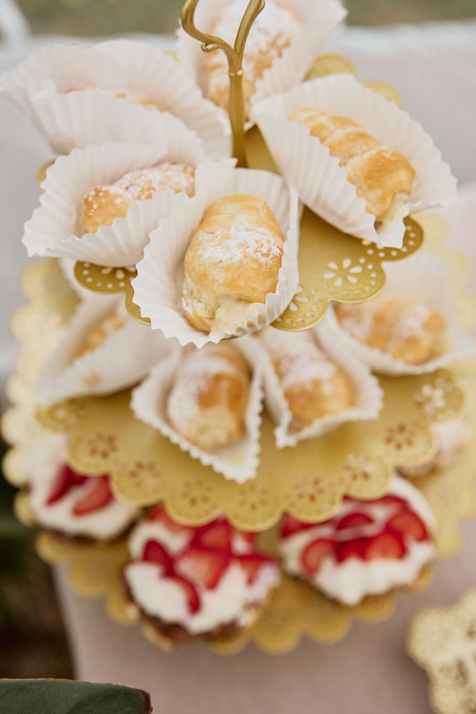 Gold tiered stand of powdered cream horns in paper liners above strawberry-topped cream pastries