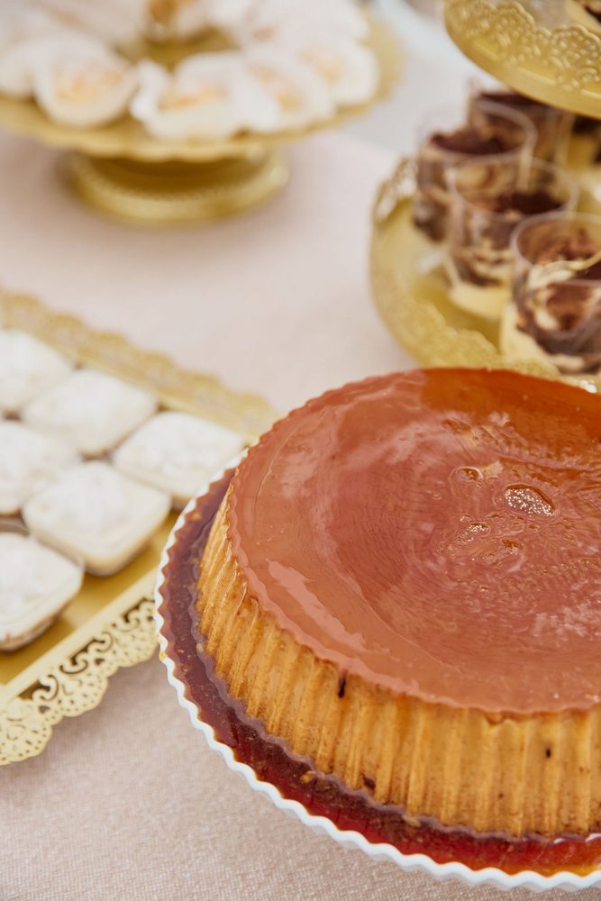 Caramel flan beside gold trays of powdered sweets and cups of layered dessert on a dessert table