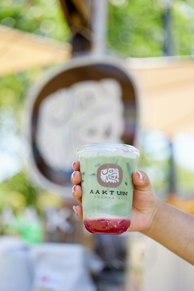 Hand holding an iced matcha drink layered over red syrup in a branded cup at an outdoor vendor