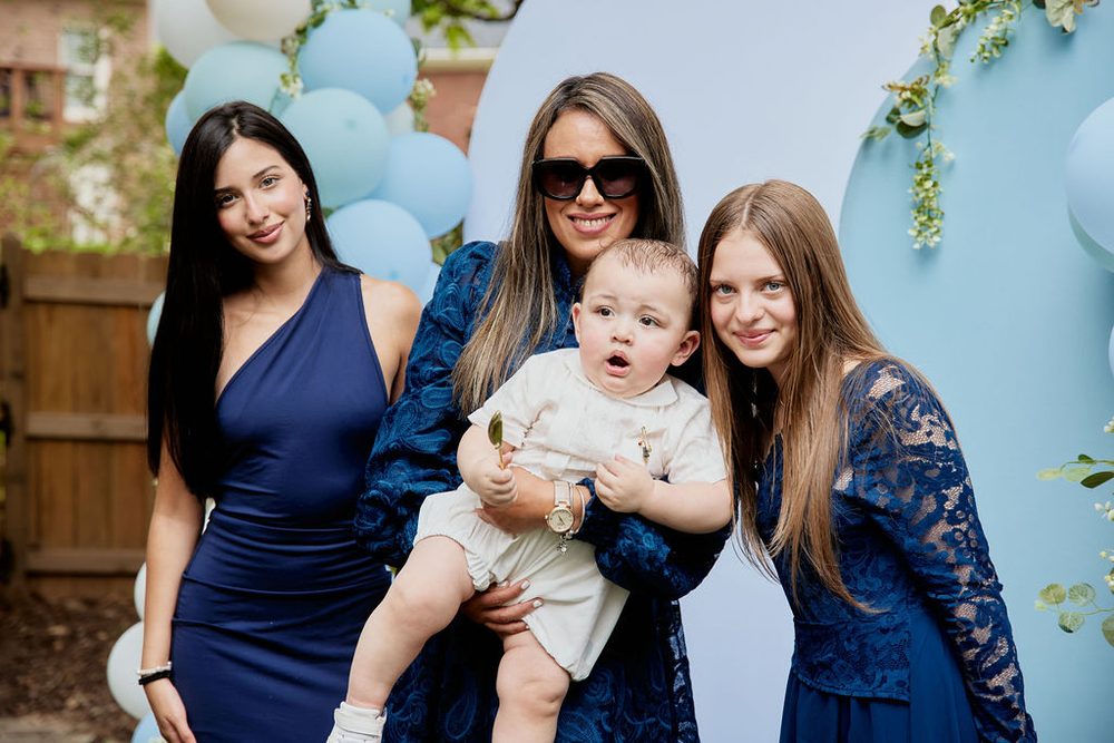 Three women in navy dresses posing with a baby in a white outfit in front of a blue balloon backdrop