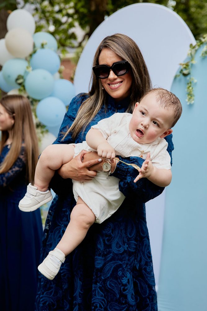 Woman in sunglasses and a navy lace dress carrying a baby in white at an outdoor celebration