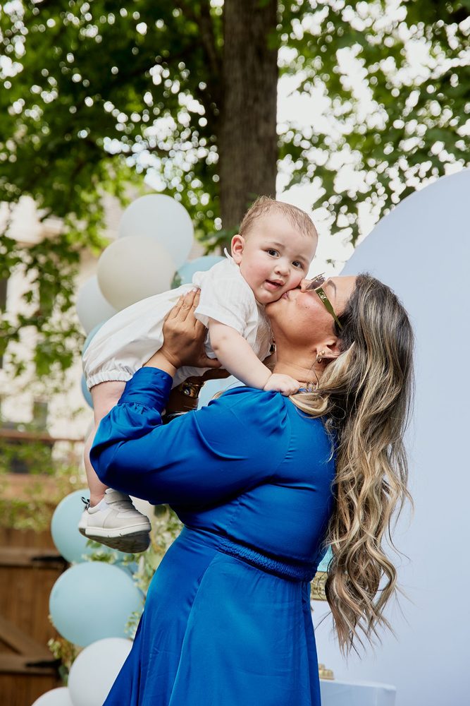 Woman with long wavy hair in a blue satin dress kissing the cheek of a baby held aloft under green trees