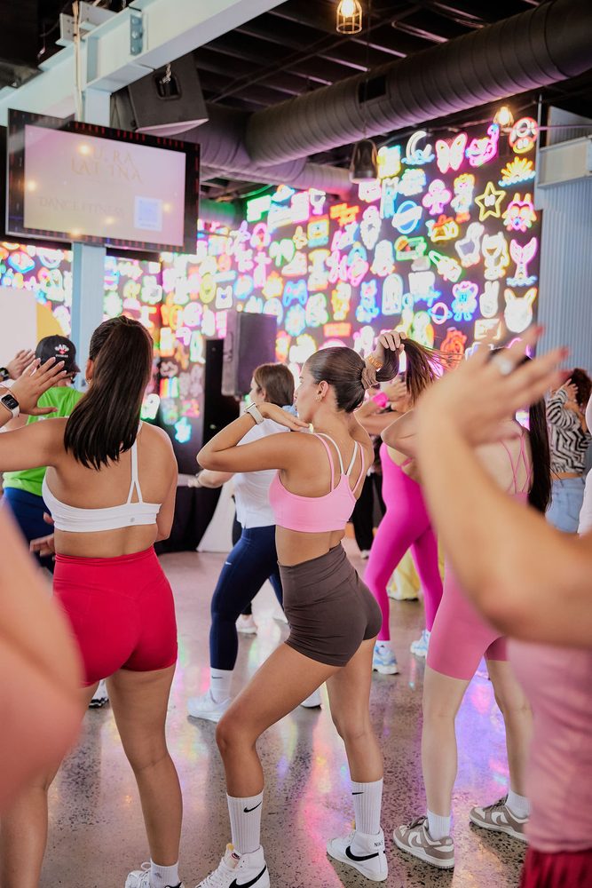 Back view of dancers in sports bras and shorts following choreography in front of a glowing neon wall