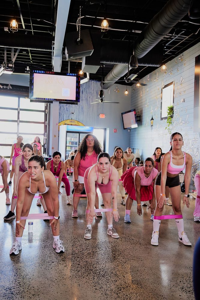 Group of women bending into squats with pink resistance bands around their legs in an industrial studio