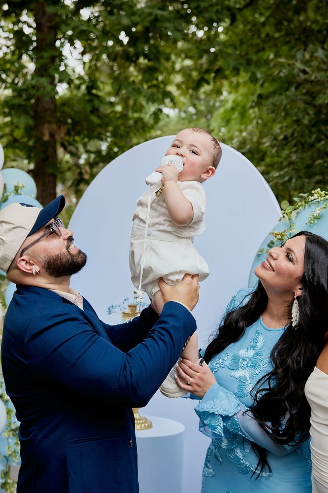 Man in a navy blazer and cap lifting a baby in white as a smiling woman in a blue lace dress looks up