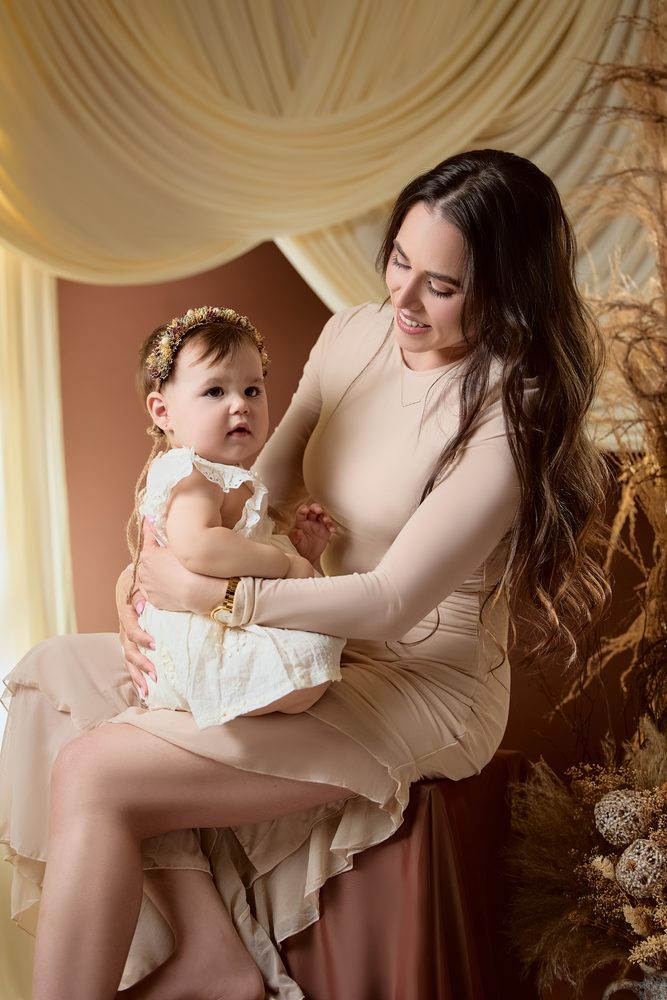 Mother in a cream dress seated with her baby girl in a floral crown on her lap, draped fabric and pampas grass behind
