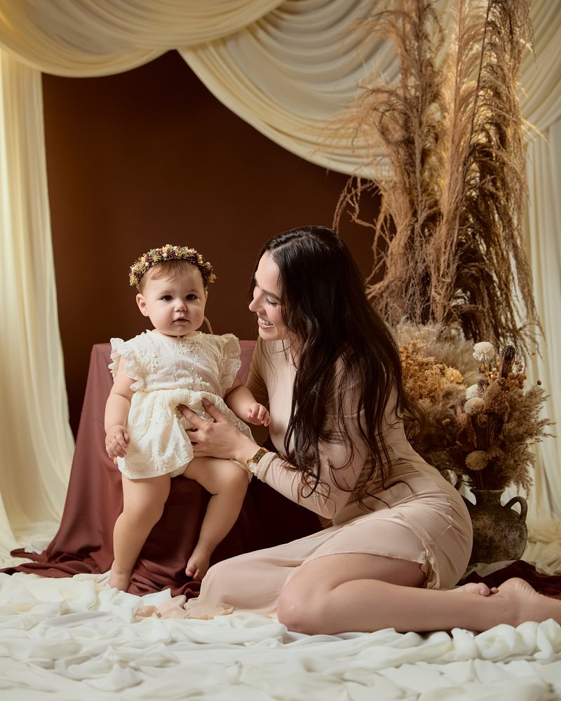 Mother sitting on white fabric steadying her standing baby girl in a lace romper and flower crown, dried pampas décor