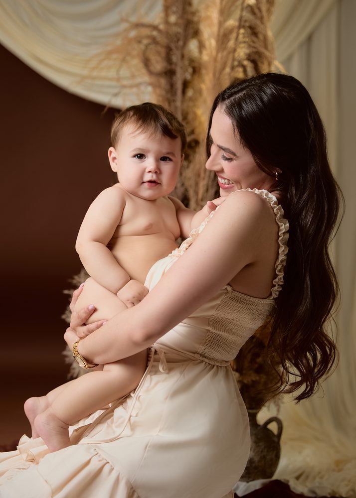 Smiling mother holding her baby up against her chest in front of tall pampas grass and cream drapes