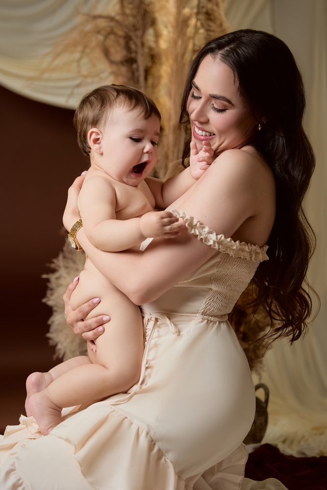 Mother laughing as her baby yawns in her arms, boho studio set with pampas grass and a brown backdrop