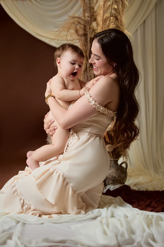 Mother kneeling on white fabric hugging her giggling baby, cream ruffled dress and clay vase beside them