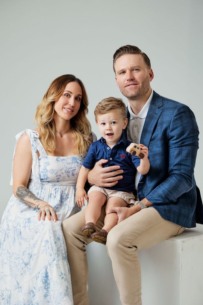 Parents seated on a white studio block with their toddler son holding a wooden toy car, light grey backdrop