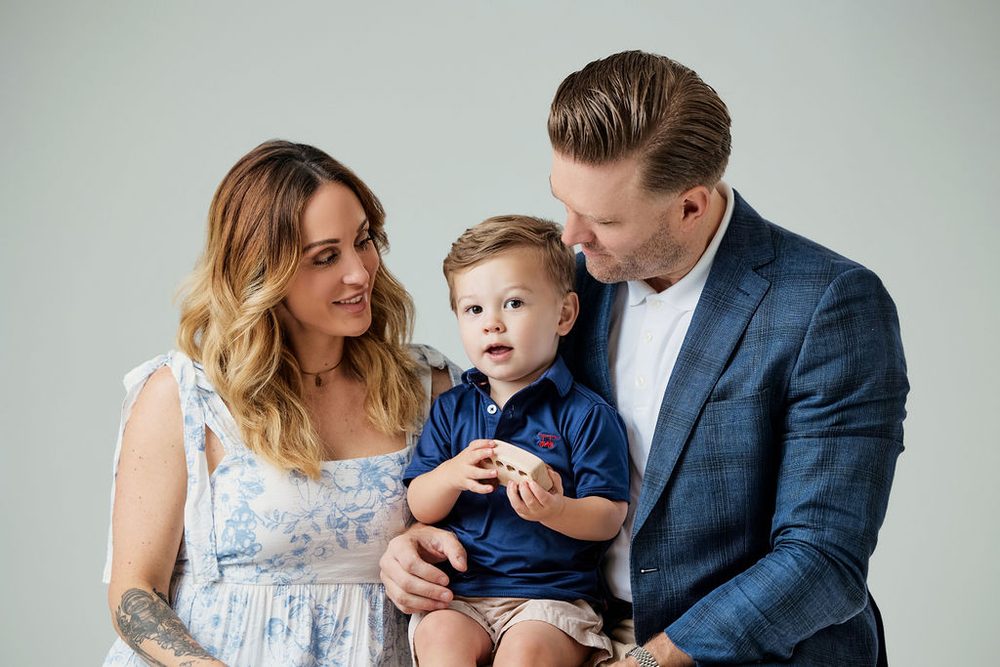 Mother and father smiling at their toddler son in a navy polo as he holds a wooden toy, grey studio background
