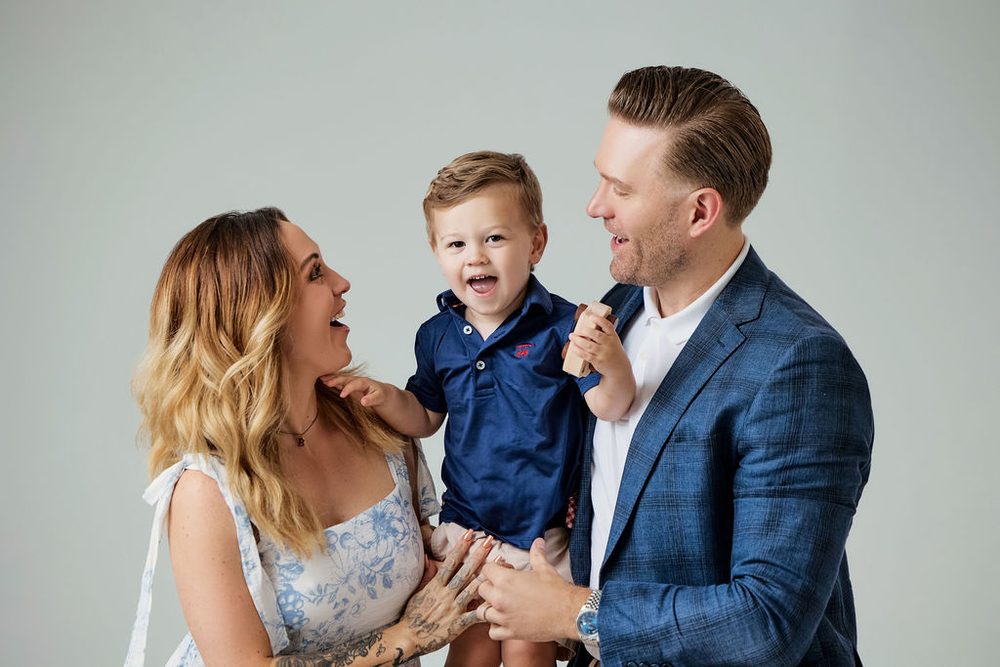Laughing toddler lifted between his mother in a blue floral dress and father in a blue blazer, grey studio